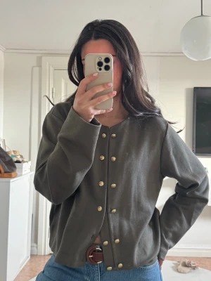 Olivgrön kofta med guldfärgade knappar - Snygg olivgrön kofta med rund halsringning och dekorativa guldfärgade knappar framtill. Rak passform och långa ärmar, perfekt att styla med jeans för en avslappnad look.