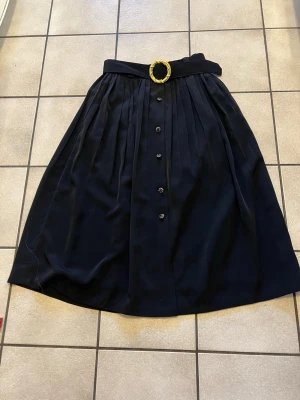 Svart långkjol med bälte och knappar storlek S/M - Stilren svart långkjol med veckad design, dekorativa knappar framtill och ett brett midjebälte med stor guldfärgad spänne. Kjolen har en klassisk och tidlös look som passar till många olika outfits.