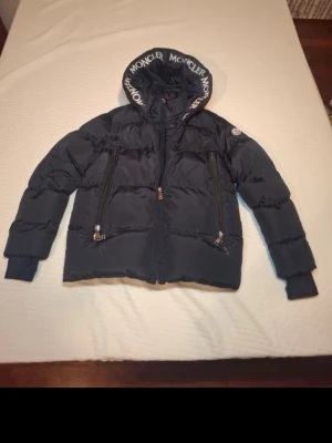 Svart dunjacka med huva från Moncler - Snygg svart dunjacka från Moncler med stor huva och tydlig logga runt huvkanten. Jackan har två snedställda dragkedjefickor framtill och quiltad design. Perfekt för kalla vinterdagar och riktigt trendig look.