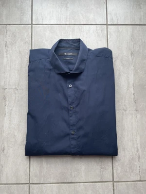 Mörkblå skjorta från Tiger of Sweden - Snygg mörkblå skjorta från Tiger of Sweden med klassisk krage och knäppning framtill. Skjortan har långa ärmar och är tillverkad i ett tunt, slätt tyg som känns bekvämt mot huden. Perfekt för en stilren look. Vid frågor och funderingar skriv gärna!