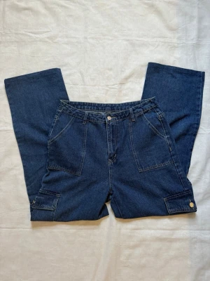 Mörkblå cargojeans med raka ben - Snygga mörkblå cargojeans i klassisk denim med raka ben och stora fickor framtill samt på benen. Jeansen har hällor för skärp och stängs med knapp och dragkedja. Perfekta för en avslappnad och trendig look.