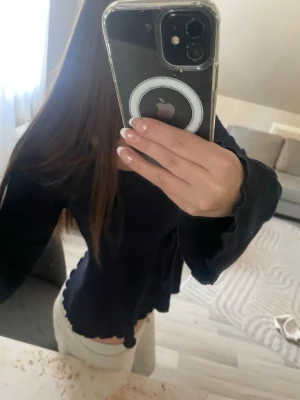 Svart långärmad topp med volangkant - Trendig svart långärmad topp med volangkant vid ärmslut och nederkant. Modellen är croppad och har en lätt utsvängd passform. Perfekt att styla med både jeans och kjol för en cool look.