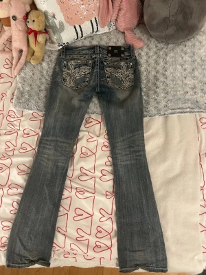 Blå bootcut jeans från Miss Me - Säljer ett par blå bootcut jeans från Miss Me med snygga broderade detaljer och bling på bakfickorna. Jeansen har låg midja och är i storlek 25. Jeansen är slim bootcut och är använda ett antal gånger. Jeansen har lite slitning längst ner.