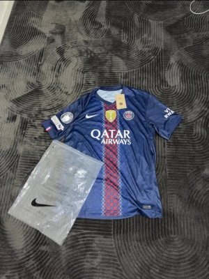 PSG matchtröja Nike blå - Snygg blå PSG matchtröja från Nike med Qatar Airways sponsortryck, klubbmärke och Nike-logga på bröstet. Röd och vit detalj längs mitten, korta ärmar och tryck på ryggen med D. Doué och nummer 14. Perfekt för fotbollsfans!