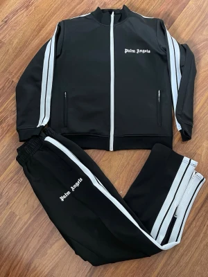 Palm Angels svart tracksuit set - Säljer ett svart tracksuit set från Palm Angels med vita ränder längs ärmar och ben. Jackan har dragkedja framtill, ståkrage och två fickor med dragkedja. Byxorna har resår i midjan och vita sidoremsor. Perfekt för chill eller streetwear vibes.