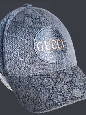 Svart keps från Gucci med GG-mönster - Snygg svart keps från Gucci med klassiskt GG-monogram över hela kepsen och en rund läderpatch framtill med guldtext. Justerbar rem baktill. Perfekt accessoar för en trendig streetstyle-look.
