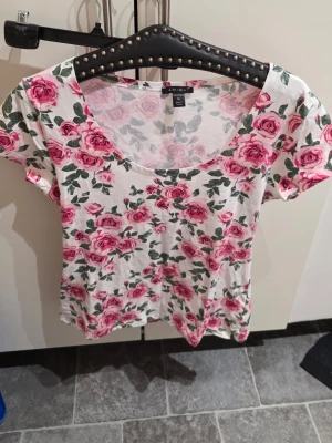 Vit topp med rosa rosor från Amisu - Supersöt vit topp från Amisu med all-over tryck av rosa rosor och gröna blad. Toppen har rund halsringning och korta ärmar. Perfekt för dig som gillar blommigt och vill ha en fräsch look till sommaren.
