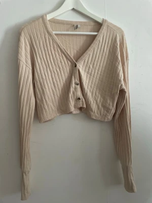 Beige kofta från NLY Trend XS - Säljer en beige cropped kofta från NLY Trend. Koftan har ribbat tyg, v-ringning och tre knappar framtill. Långa ärmar med breda muddar och en avslappnad passform.