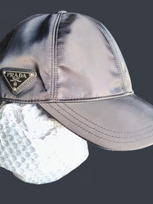 Svart keps från Prada i re- nylon - Snygg svart keps från Prada i glansigt nylonmaterial. Klassisk böjd skärm och detaljerade sömmar. På sidan sitter Pradas ikoniska metalltriangel med logga. Perfekt accessoar för en trendig streetwear-look.