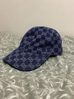 Blå denimkeps från Gucci - Snygg blå keps från Gucci i denim med det klassiska GG-mönstret över hela kepsen. Justerbar rem baktill i läder och gulddetalj. Perfekt accessoar för att lyfta din streetwear-look.