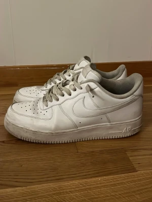 Nike Air Force 1 vita sneakers - Klassiska vita Nike Air Force 1 sneakers med lågt skaft. Skorna har perforerad tå, vita skosnören och den ikoniska swoosh-loggan på sidan. Tillverkade i läder med en robust sula där det står 'AIR' på sidan. Perfekta för en clean och tidlös streetstil.