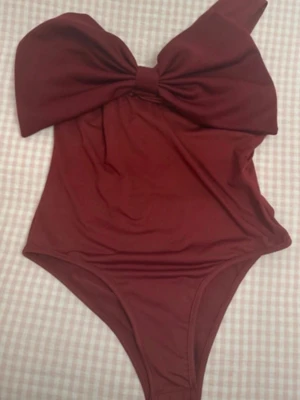 Vinröd body med stor rosett - Supersnygg vinröd body med en stor dekorativ rosett framtill. Asymmetrisk axelrem ger en trendig look. Perfekt för dig som vill sticka ut på en utekväll. Materialet känns mjukt och stretchigt.