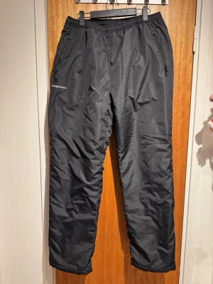 Svarta täckbyxor från Bagheera - Svarta täckbyxor från Bagheera med elastisk midja och dragkedjefickor. Perfekta för kalla dagar och vinteraktiviteter. Tillverkade i syntetmaterial som håller dig varm och torr. 