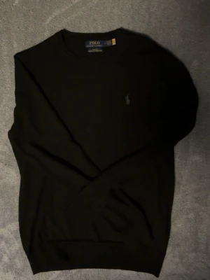 Svart merinoulltröja Polo Ralph Lauren - Klassisk svart stickad tröja från Polo Ralph Lauren i slim fit-modell. Tillverkad i tvättbar merinoull och har den ikoniska broderade loggan på bröstet. Perfekt basplagg med rund hals och långa ärmar.