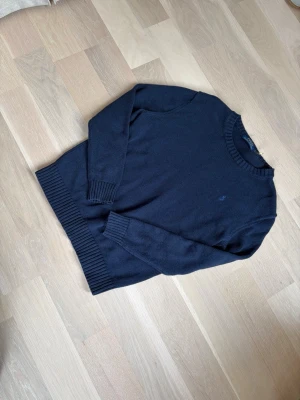 Mörkblå stickad tröja Polo Ralph Lauren - Mörkblå stickad tröja från Polo Ralph Lauren med rund halsringning och diskret broderad logga på bröstet. Tröjan har långa ärmar och ribbstickade muddar vid ärmslut och nederkant. Perfekt för en stilren och klassisk look.