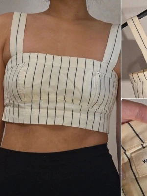 Randig linnetopp från Wood Wood - Säljer en snygg cremevit linnetopp från Wood Wood med svarta vertikala ränder. Toppen har breda axelband, croppad passform och dragkedja i sidan. Perfekt för dig som gillar minimalistisk och stilren design. Finns solkrämsfläckar vid insidan