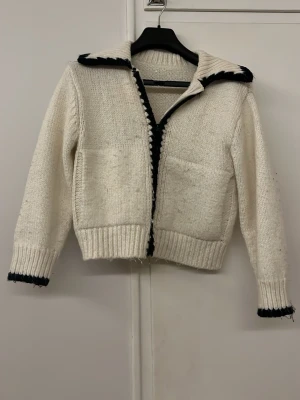 Vit stickad kofta  - Säljer en vit stickad kofta med svarta kanter längs krage, ärmslut och öppning. Koftan har två fickor framtill, ribbstickade muddar och en bred krage. Perfekt för lager-på-lager och en chill look. Den är liten i armarna så skulle typ säga att den passar till yngre