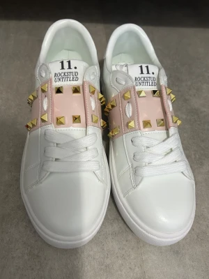Valentino Rockstud sneakers vit/rosa - Säljer ett par Valentino Garavani Rockstud sneakers i vitt skinn med ljusrosa detaljer och guldiga nitar. Klassisk rund tå, snörning och platt sula. Ikoniska pyramidnitar längs sidorna och bak på hälen. Kommer med originalkartong och dustbag. Passar dig som vill ha en lyxig stil nu i det nya året.