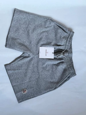 Grå shorts från Moncler med snörning - Snygga grå shorts från Moncler med elastisk midja och snörning. Tillverkade i mjuk bomull och har Moncler-logga broderad på ena benet. Perfekta för chill dagar eller träning. Klassisk passform och stilren design.