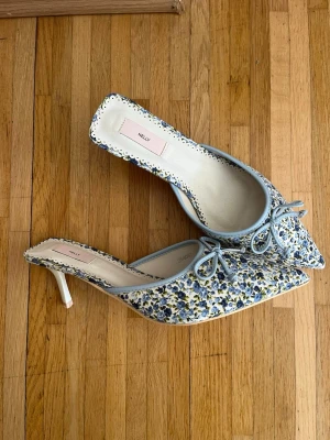 Blommiga kitten heels med rosett från Nelly - Säljer dessa populära pumps från Nelly med blommigt mönster i blått, gult och vitt. Skorna har en söt rosettdetalj framtill och ljusblå kant. De är aldrig använda.