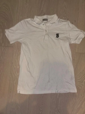 Vit pikétröja från Burberry - Vit pikétröja från Burberry med klassisk krage och knappar framtill. Tröjan har korta ärmar och en broderad svart logotyp på bröstet. Perfekt för en clean och stilren look.