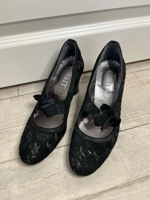 Svarta spetsiga pumps med rosett - Snygga svarta pumps i storlek 36 men passar 37 från Scorett med rundad tå och dekorativ rosett framtill. Skorna har ett elegant spetsmönster och en klassisk klack. Materialet är en kombination av textil och spets som ger en lyxig känsla. Perfekta för dig som vill ha en unik och stilren look.