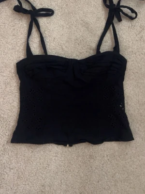 Svart topp med knytband - Svart topp med knytbara axelband och dekorativa broderade detaljer på sidorna. Toppen har en dragkedja i ryggen och är croppad i modellen. Perfekt för dig som vill ha en trendig och stilren look. Toppen är ifrån Zara. Toppen har ett litet hål precis vid dragkedjan, som du ser på bild 5. Påverkar dock inte själva dragkedjan.
