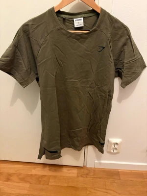 Olivgrön t-shirt från Gymshark - Olivgrön t-shirt från Gymshark med rund halsringning och korta ärmar. Diskret Gymshark-logga på bröstet. Perfekt för träning eller gymmet. Tillverkad i ett lätt och stretchigt material som andas.