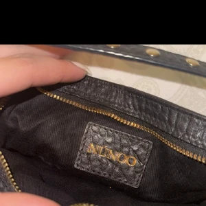 Svart axelväska från Nunoo - Snygg svart axelväska från Nunoo i strukturerat läder med guldfärgade dragkedjor och detaljer. Väskan har två små fickor framtill och justerbar axelrem. Perfekt för dig som vill ha en trendig och praktisk väska med coola detaljer.