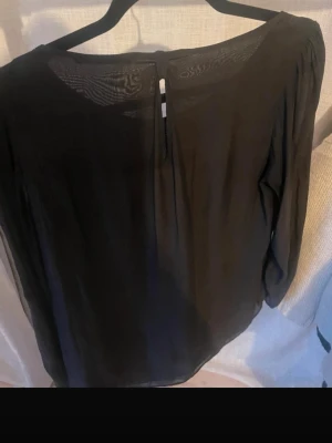 Svart transparent blus med rund hals från esprit  - Snygg svart blus i transparent material med rund halsringning och knappdetalj bak. Blusen har långa, luftiga ärmar och ett lätt, flödande tyg som ger en avslappnad men chic look. Perfekt att styla med jeans eller kjol för en trendig outfit.