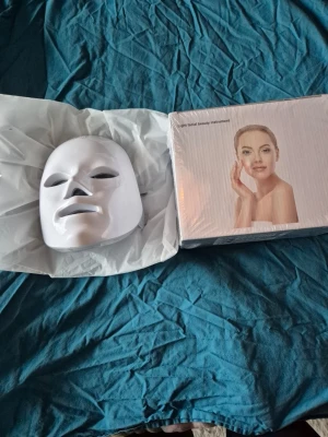 LED ansiktsmask för hudvård - LED ansiktsmask i vit plast med justerbara gråa band. Masken har flera små LED-lampor på insidan och används för ljusbehandling av huden. Kommer i originalförpackning med bildinstruktioner på kartongen.