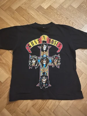 Guns N' Roses t-shirt - Svart t-shirt med stort färgglatt Guns N' Roses-tryck på framsidan. Motivet har ett kors med bandmedlemmarnas döskalleporträtt och loggan i gult och rött ovanför. Klassisk passform och rund hals. Perfekt för dig som gillar rock och bandmerch. Väldigt fint skick köpt secondhand