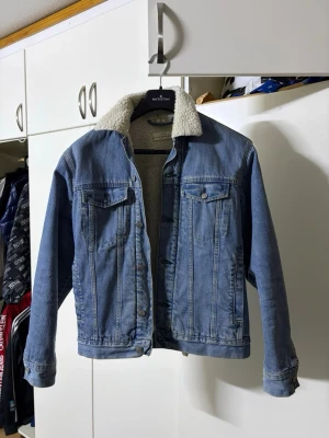 Blå jeansjacka med teddyfoder - Säljer en klassisk blå jeansjacka från BATTINNI med vitt teddyfoder i kragen och på insidan. Jackan har två bröstfickor med lock och knappar samt knappstängning framtill. Perfekt för dig som vill ha en tidlös och trendig look.