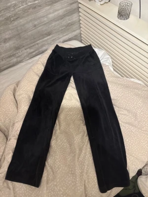 Svarta mjukisbyxor Juicy Couture - Säljer ett par klassiska svarta mjukisbyxor från Juicy Couture i mjuk velour. Byxorna har raka ben, dragsko i midjan och diskret broderad logga på fickan. Perfekta för chill dagar eller när du vill vara extra bekväm.