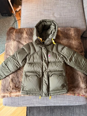 Grön dunjacka från Fjällräven - HELT NY. Snygg grön dunjacka från Fjällräven med stor huva och gula detaljer på snörningen. Jackan har två stora fickor framtill med lock och tryckknappar samt klassisk Fjällräven-logga på bröstet. Perfekt för kalla vinterdagar och riktigt varm. Färgen heter deep forest. Nypris: 7499kr 