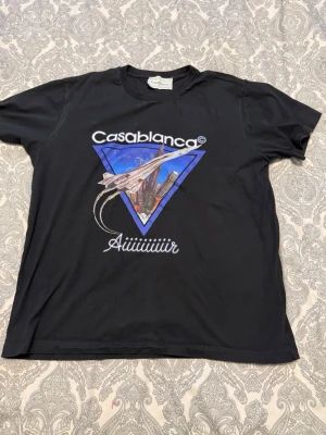Svart Casablanca t-shirt med tryck - Svart t-shirt från Casablanca med stort grafiskt tryck framtill som visar ett flygplan och stadssilhuett i blå triangel. Texten 'Casablanca' och 'Aiiiiiiiir' syns på bröstet. Klassisk passform och rund hals. Perfekt för dig som gillar streetwear och coola prints.