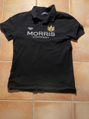 Svart Morris Company piké - Snygg svart piké från Morris Company med vit brodyrtext och guldfärgat emblem på bröstet. Klassisk pikékrage och korta ärmar. Tillverkad i mjuk bomull för skön känsla och stilren look.