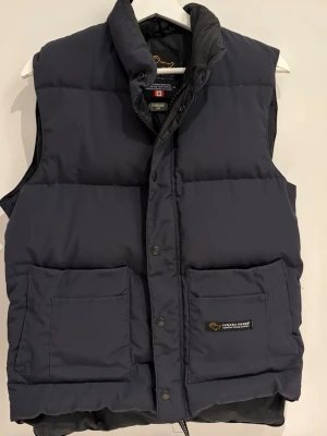 Mörkblå dunväst från Canada Goose - Snygg mörkblå dunväst från Canada Goose med hög krage och två stora framfickor. Västen har en klassisk puffer-design och stängs med både dragkedja och tryckknappar. Perfekt för lager-på-lager och streetstyle.
