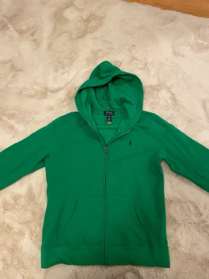 Grön hoodie från Polo Ralph Lauren - Grön hoodie med dragkedja från Polo Ralph Lauren. Klassisk modell med huva, två fickor framtill och broderad logga på bröstet. Perfekt för en avslappnad stil och enkel att matcha med jeans eller joggers. Passar som storlek S. Pris går att diskutera. Hör av dig vid minsta lilla fundering!