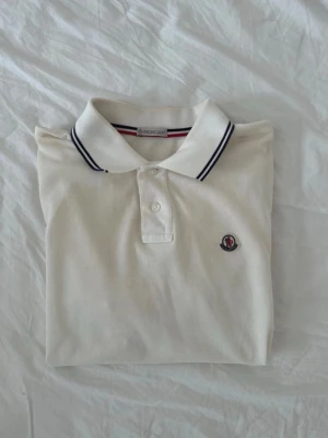 Vit pikétröja från Moncler - Klassisk vit pikétröja från Moncler med broderad logga på bröstet och krage med svarta ränder. Tröjan har korta ärmar och knäppning framtill. 