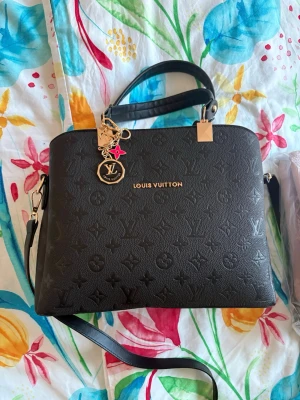 Svart Louis Vuitton handväska - Snygg svart handväska från Louis Vuitton i präglat läder med klassiskt LV-monogram. Väskan har både axelrem och handtag, guldfärgade detaljer samt en dekorativ nyckelring med LV-logga och rosa stjärna. Invändigt finns flera fack med dragkedja för smart organisering.