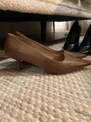 Bruna pumps med låg klack från HM - Snygga bruna pumps med spetsig tå och låg klack. Skorna har ett stilrent ormmönstrat yttre i konstläder och en klassisk siluett