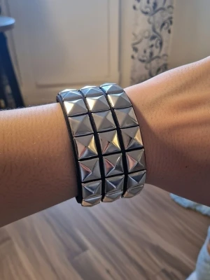Silver, svart punkarmband - Ett svart punkarmband med tre rader av silverfärgade pyramidformade nitar. Armbandet stängs med tryckknappar. PU läder. Storlek 22 cm långt. Bredd 4,6 cm.