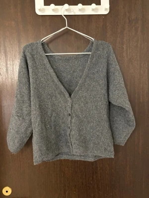 Grå mjuk kofta med v-ringning trekvartsärmad - En enkel och mysig grå kofta med v-ringning och knappar framtill. Koftan har trekvartsärm och är gjord i ett mjukt, stickat material som känns skönt mot huden. Perfekt att slänga på sig när det är lite kyligare ute. Aldrig använd.