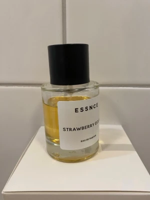ESSNCE Strawberry Eclair parfym - En stilren parfymflaska från ESSNCE med doften Strawberry Eclair. Flaskan är cylindrisk i genomskinligt glas med svart rund kork och vit etikett med svart text. Innehållet är bärnstensfärgat och ger ett fräscht och modernt intryck. Mängd enl bilder på flaskan