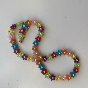 Färgglatt halsband med stjärnpärlor - Superfint halsband med stjärnformade pärlor i olika färger som rosa, blå, grön, lila, orange och röd. Halsbandet har en guldfärgad kedja och är perfekt för att ge din outfit en lekfull touch. Materialet är plast och metall.