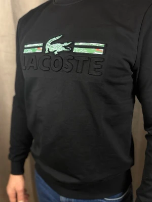 Svart långärmad tröja från Lacoste - Svart långärmad tröja från Lacoste med stort broderat logotyptryck och gröna detaljer framtill. Tröjan har rund halsringning och ribbade muddar vid ärmslut och nederkant. Perfekt för en stilren och sportig look.