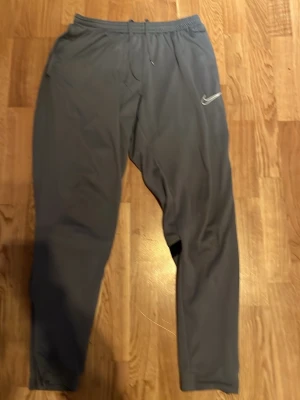 Grå Nike mjukisbyxor med logga Academy - Snygga grå mjukisbyxor från Nike med vit logga på ena benet. Byxorna har resår i midjan med dragsko och svarta detaljer baktill vid knäna. Perfekta för chill eller träning, tillverkade i mjukt och stretchigt material.