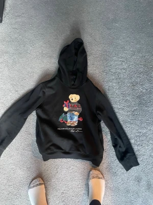 Svart hoodie Polo Ralph Lauren björn - Svart hoodie från Polo Ralph Lauren med ikonisk björn-brodyr på bröstet. Hoodien har långa ärmar, huva och ribbade muddar. Perfekt för dig som gillar streetstyle och vill ha något klassiskt men ändå lekfullt.