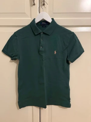 Grön pikétröja från Ralph Lauren - Klassisk grön pikétröja från Polo Ralph Lauren med korta ärmar och slim fit-passform. Tröjan har krage, knappar framtill och den ikoniska broderade loggan på bröstet. Perfekt till både jeans och shorts.